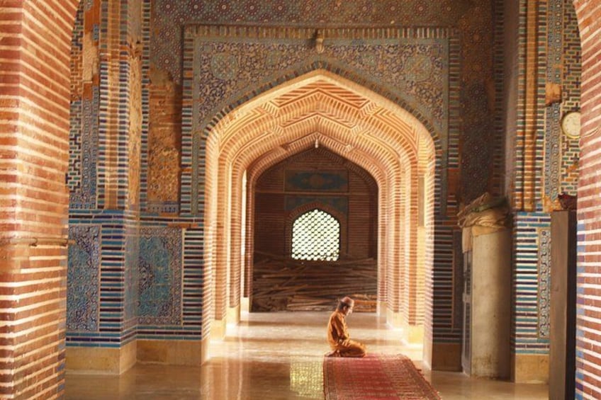 Une personne en prière, dans un intérieur richement décoré, orné de motifs complexes en mosaïque et doté d’une porte en arc de cercle. 1