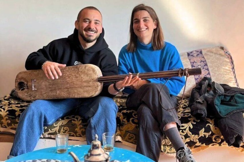 Two people sitting on a couch with a guitar and a teapot on a table. 3