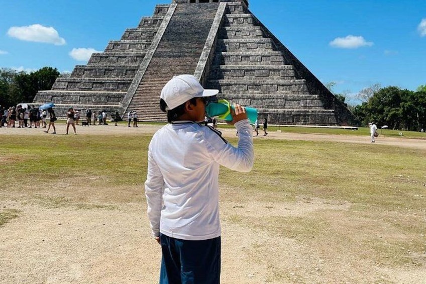 Een persoon in een wit shirt en pet drinkt uit een groene fles, staand voor een grote piramide. 2