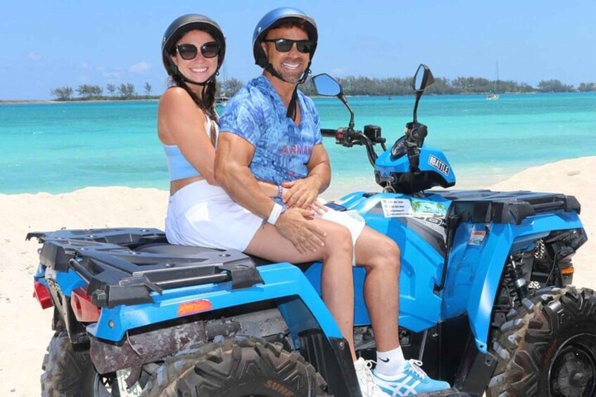 Two people on a blue ATV on a sandy beach with turquoise water and a clear sky. 5