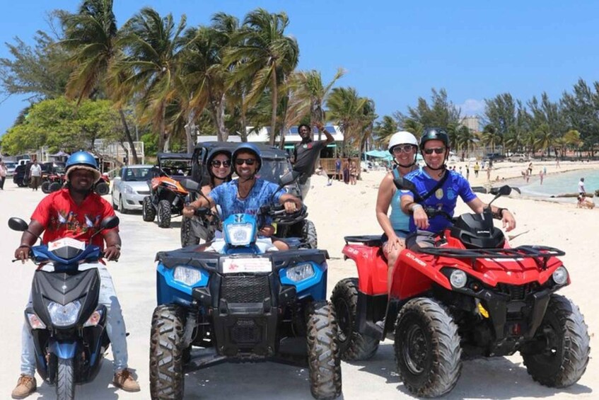 Four people on all-terrain vehicles on a beach with palm trees and parked cars in the background. 1