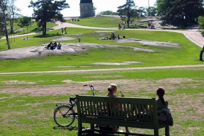 A park with a green bench, a bicycle, and people sitting on rocks. 3