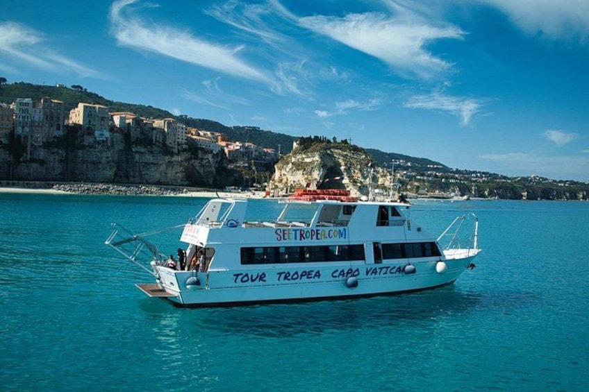 A white boat named 'TOUR TROPEA CAPO VATICAN' with 'SETROPEAL.COM' on its side, sailing on turquoise waters with a coastal town and cliffs in the background. 2