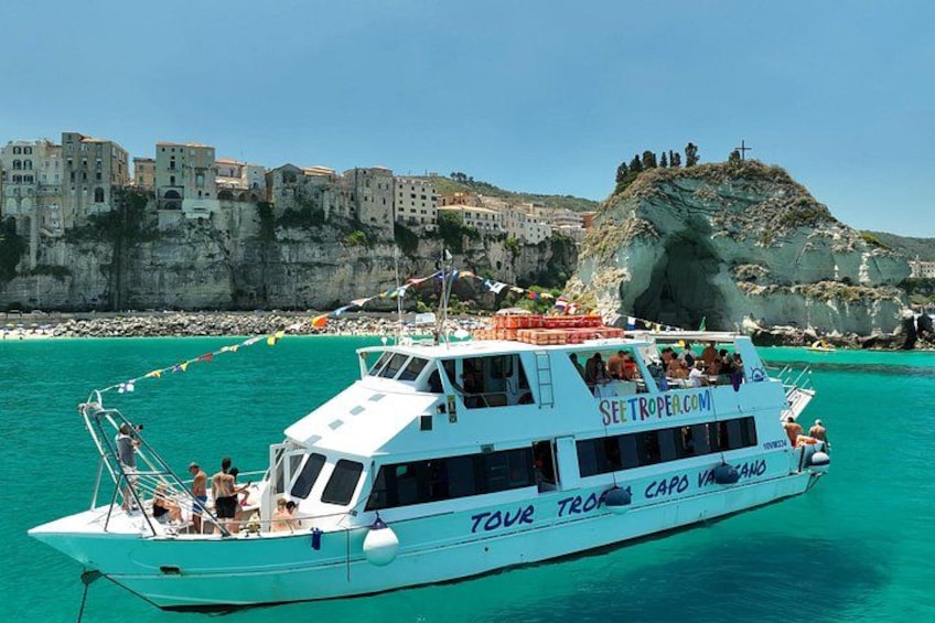 A white boat with 'SEETROPE.COM' and 'TOUR TROPE CAPO VANO' on its side, sailing in turquoise waters with a rocky coastline and buildings in the background. 1