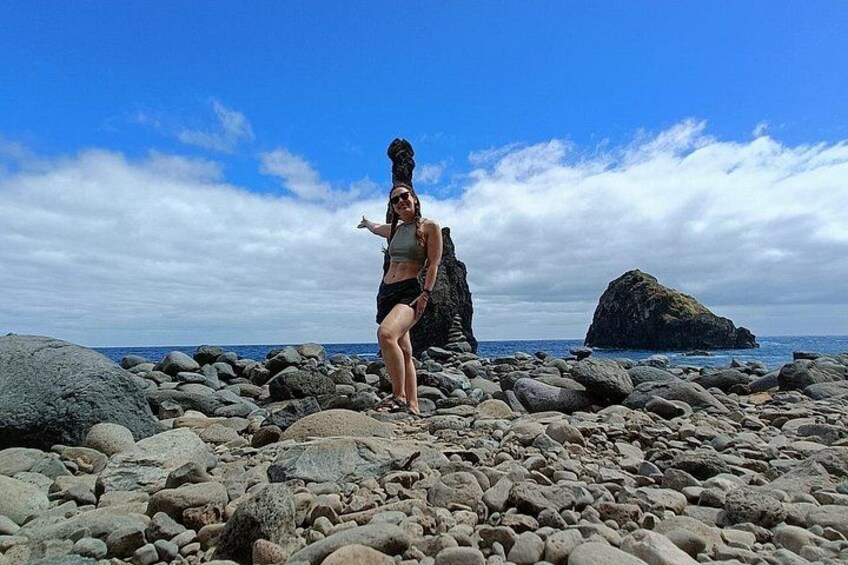 A person standing on a rocky beach with large boulders, a clear sky, and a distant rock formation. 4