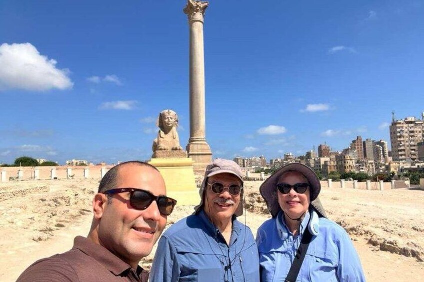 Three people standing in a sandy area with a large column and a statue in the background. 1
