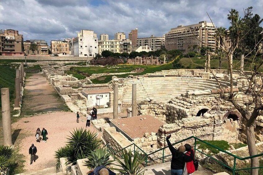 Ancient ruins with a theater, visitors, and modern buildings in the background. 2