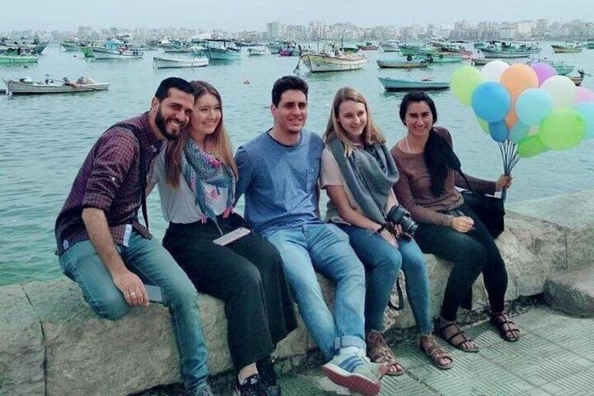 A group of people sitting on a stone ledge by a body of water with boats in the background. 3