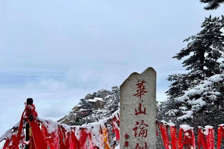 Paisaje nevado de una montaña con una persona vestida de rojo escalando una pendiente cubierta de nieve, un monumento de piedra con caracteres chinos y árboles cubiertos de nieve. 1