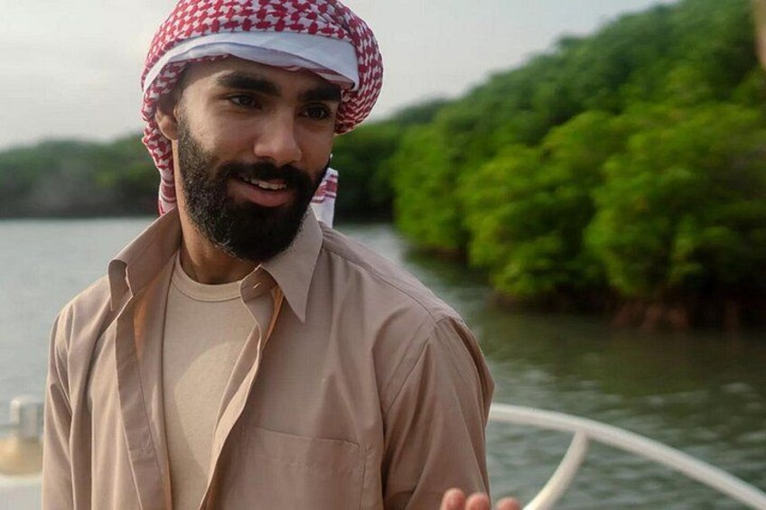 A person wearing a traditional headscarf and a light-colored shirt, standing by a body of water with greenery in the background. 1