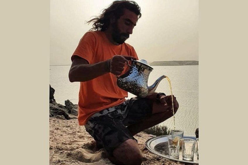 En person hælder væske fra et metaltermokande i et glas på en strand. 3