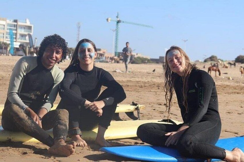 Tre personer sidder på surfboards på en sandstrand med en kran og bygninger i baggrunden. 4