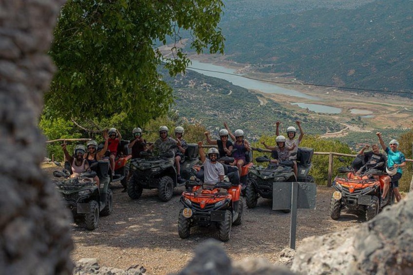 A group of people on quad bikes on a dirt path with a scenic view of a valley and a lake in the background. 1