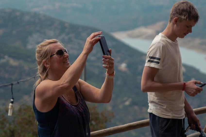 A person taking a selfie with a smartphone, another person looking at a smartphone, and a scenic mountainous landscape in the background. 5