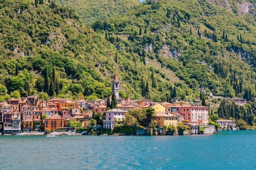 A coastal town with colorful buildings, a prominent tower, and a backdrop of lush green hills. 1