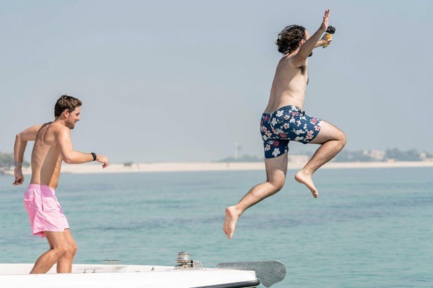 A person jumping off a boat into the water, another person on the boat, and a beach with sand and water in the background. 2