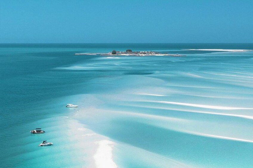 A clear blue sea with a sandy beach, a small island with buildings, and three boats. 1