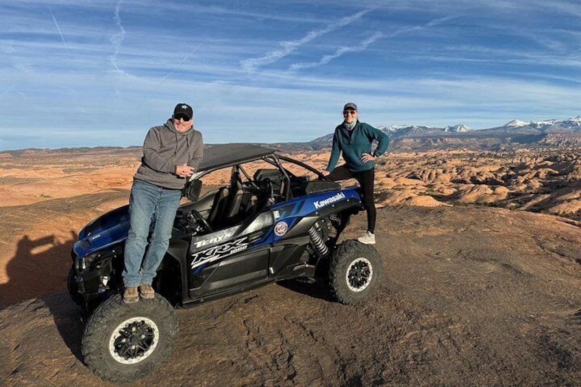 Deux personnes se tiennent à côté d’un véhicule tout-terrain bleu sur un terrain rocheux, avec un paysage montagneux en arrière-plan. 2