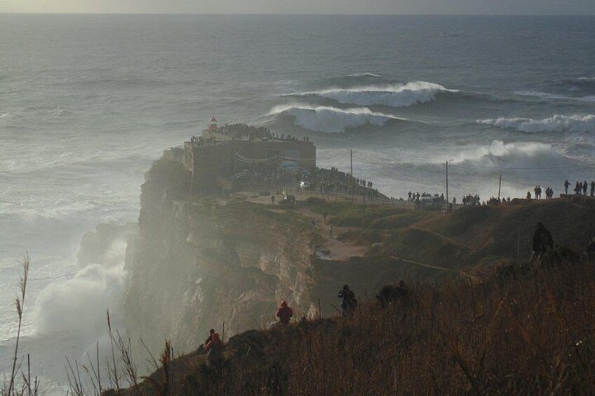 Un paysage côtier avec une structure fortifiée, des vagues qui s’écrasent sur le rivage et des personnes qui observent la scène. 5