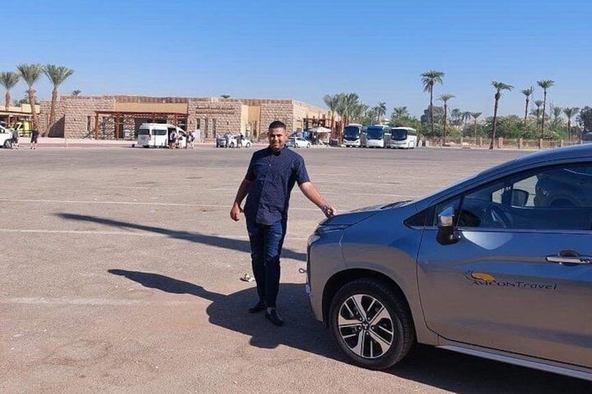 A man standing next to a car in a parking lot with a building and palm trees in the background. 1