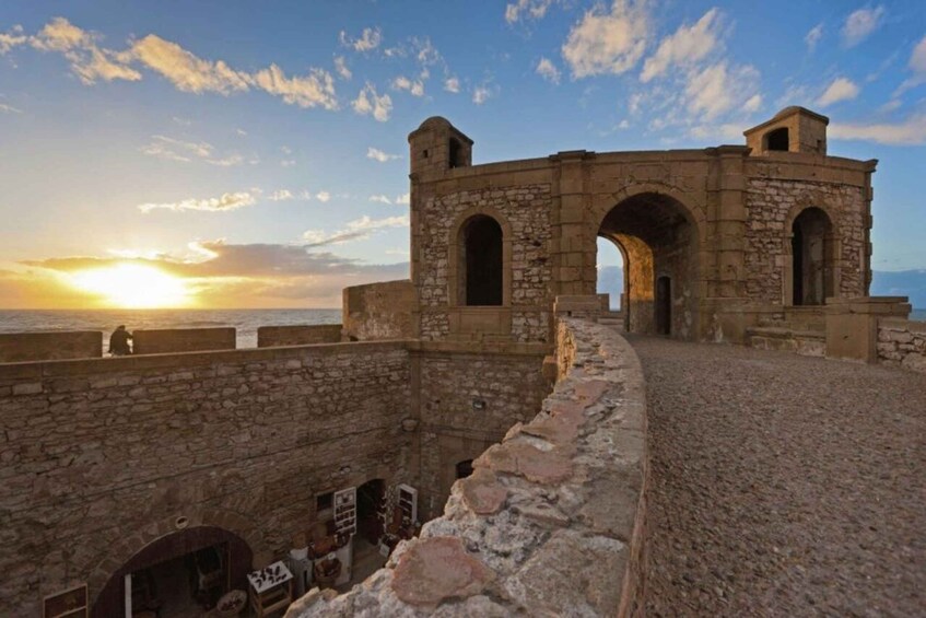 A stone fortress with arched gateways overlooking the sea at sunset. 1