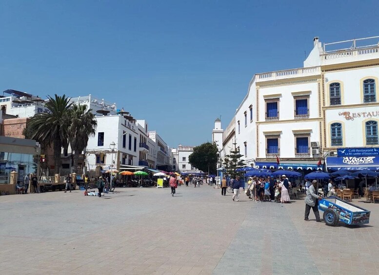 A bustling square with people walking, palm trees, and buildings with cafes. 3