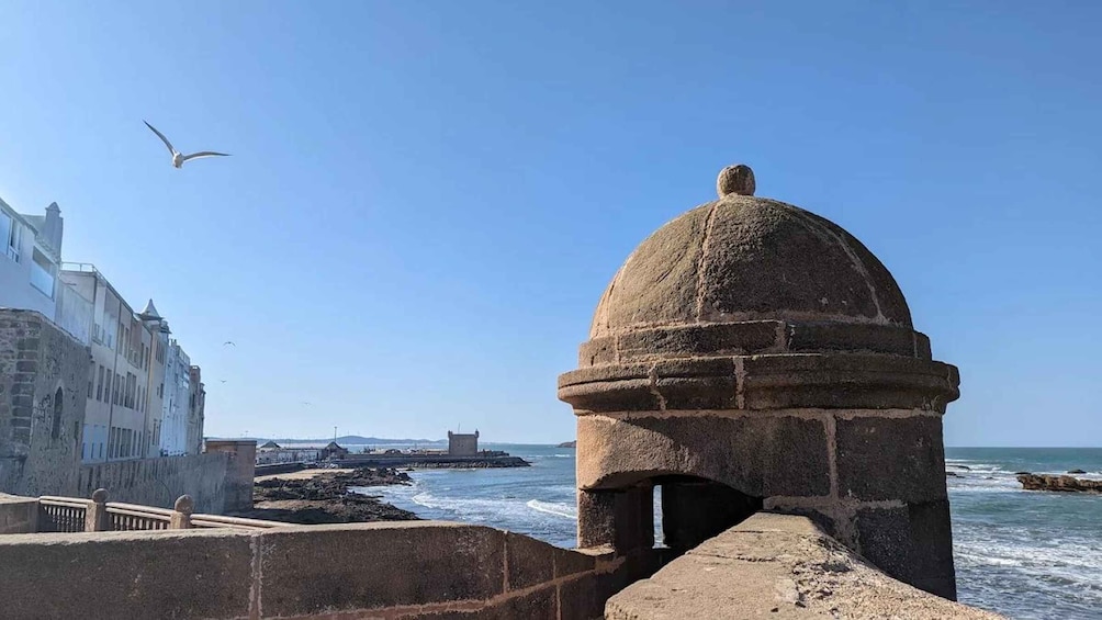 A coastal scene with a stone structure, a seagull flying, and buildings in the background. 5
