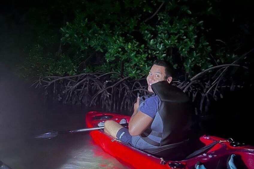 A person in a red kayak with a life jacket, paddling through a mangrove area at night. 4
