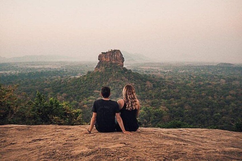 Two people sitting on a hill overlooking a landscape with a prominent rock formation. 3