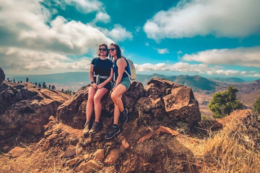 Two people sitting on a rocky outcrop with a mountainous landscape in the background. 4