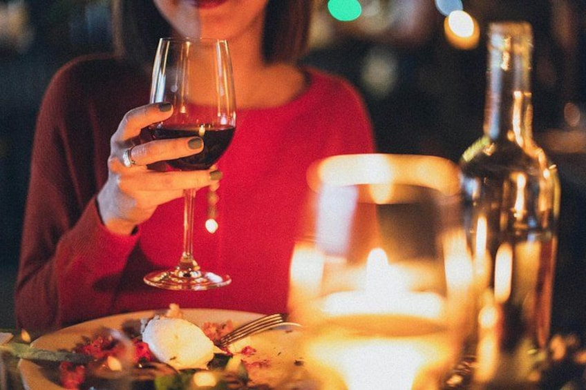 A person holding a wine glass, a candle, and a bottle on a table with a plate of food. 1