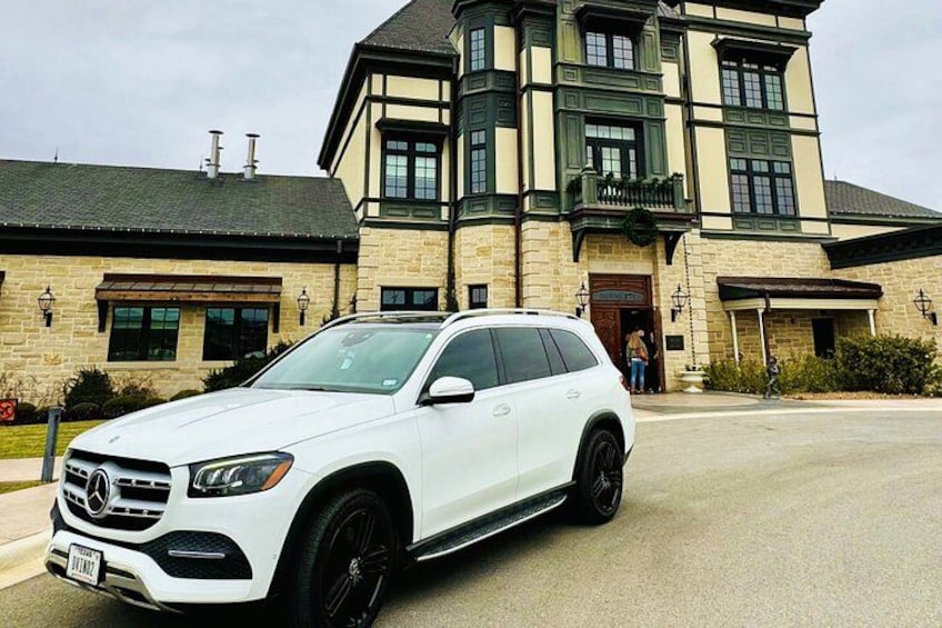 Un SUV bianco parcheggiato di fronte a una grande casa a più piani con facciata in pietra e un balcone verde. 1