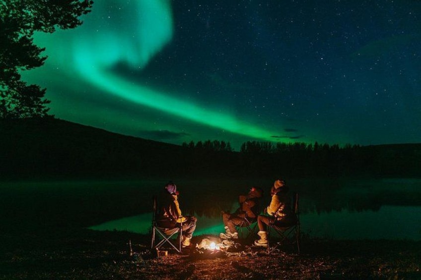 Three people sitting by a campfire under a night sky with the aurora borealis. 1