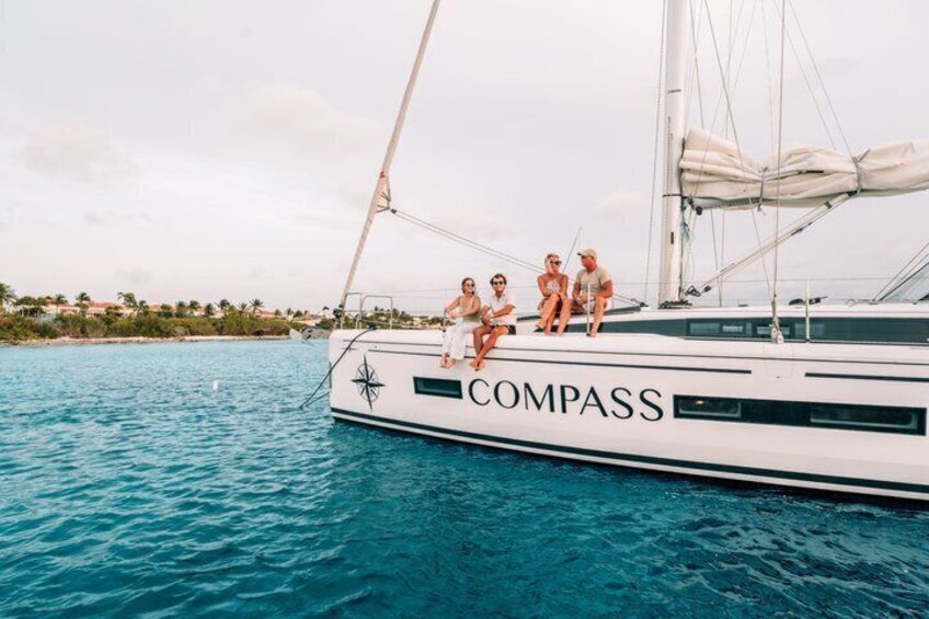 A white yacht named 'COMPASS' with people sitting on the deck, surrounded by turquoise water and a coastal landscape. 4