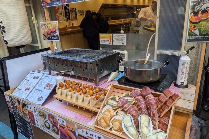 Un puesto de comida con una parrilla, una olla de sopa y distintos alimentos a la vista. 5