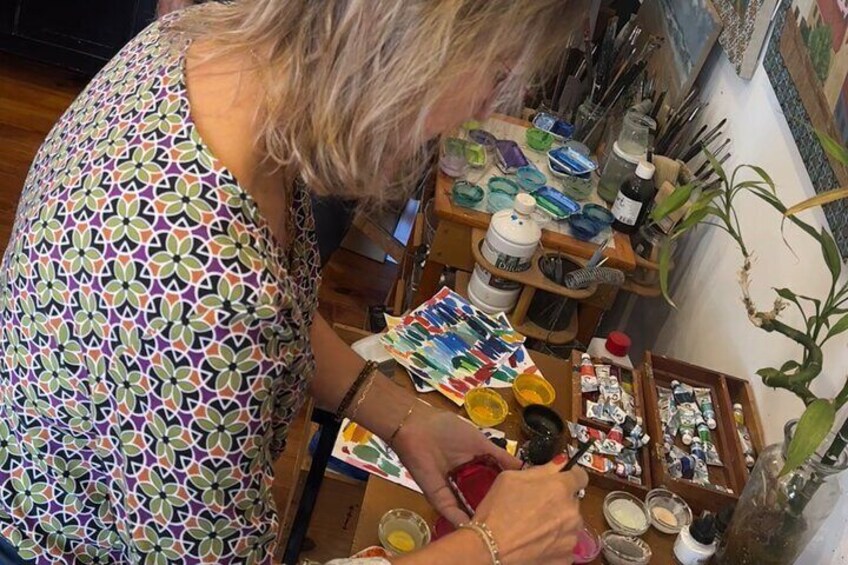 Iemand schildert aan een tafel, omringd door verschillende kunstbenodigdheden en een werk in uitvoering. 5