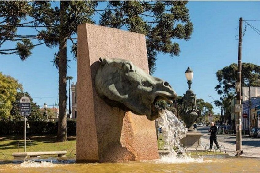A stone fountain with a horse head sculpture, surrounded by trees and a street lamp. 4