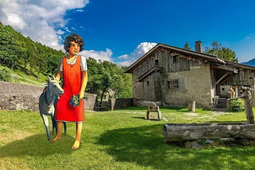 A person in a red dress standing in a grassy field with a cow, a rustic wooden house, and a stone wall in the background. 2