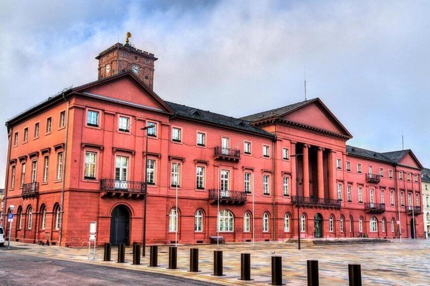 A red brick building with multiple windows and balconies, a clock tower, and a paved square in front. 4