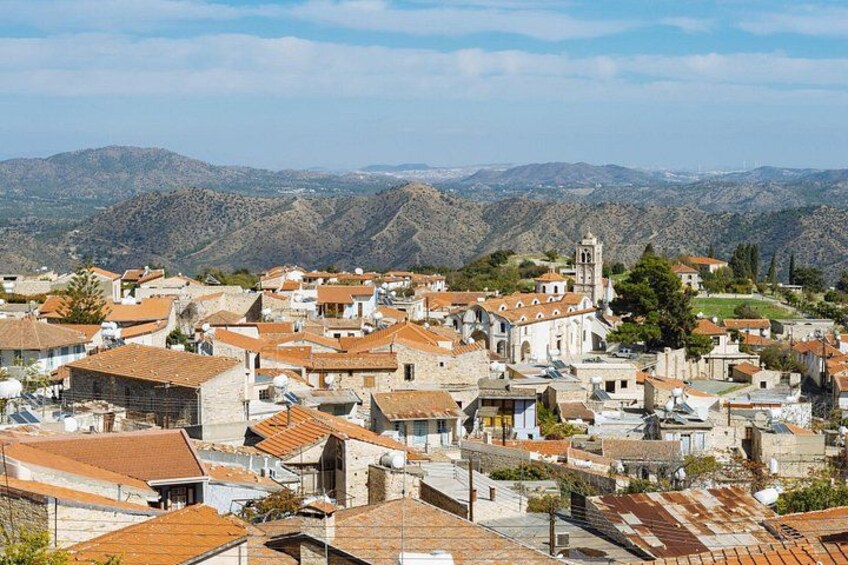 A town with terracotta-roofed buildings, a prominent church with a bell tower, and a mountainous backdrop. 2