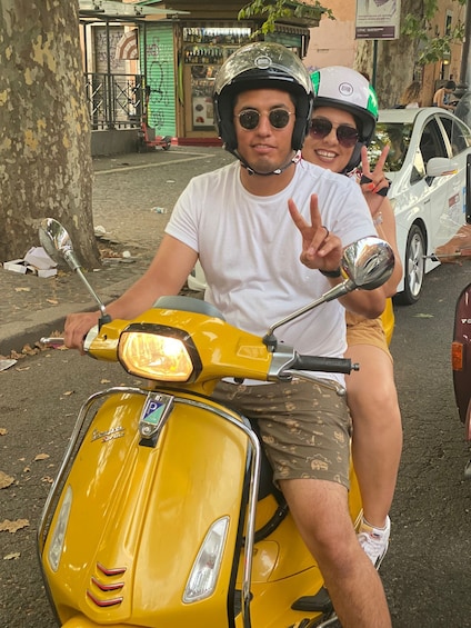 Keltainen Vespa-skootteri, jonka päällä istuu henkilö, toinen henkilö valkoisella skootterilla ja valkoinen auto, joka on pysäköity lähelle. 1