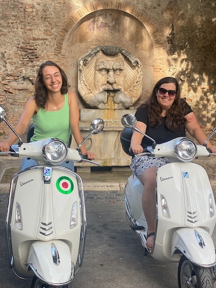 Kaksi ihmistä ajaa vanhoilla Vespa-moottoriskoottereilla kivestä tehdyn kaarimaisen portin edessä, jossa on leijonan kasvojen kuva. 3