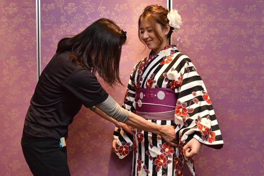 En person bär en kimono med blommönster, en annan person hjälper till med att sätta på kimonon, och bakgrunden har ett lila mönster. 1