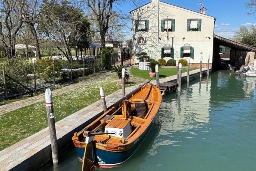 Ein schmaler Kanal mit einem hölzernen Boot, das an einem hölzernen Steg anliegt, ein Gebäude mit grünen Fensterläden und ein klarer Himmel. 4