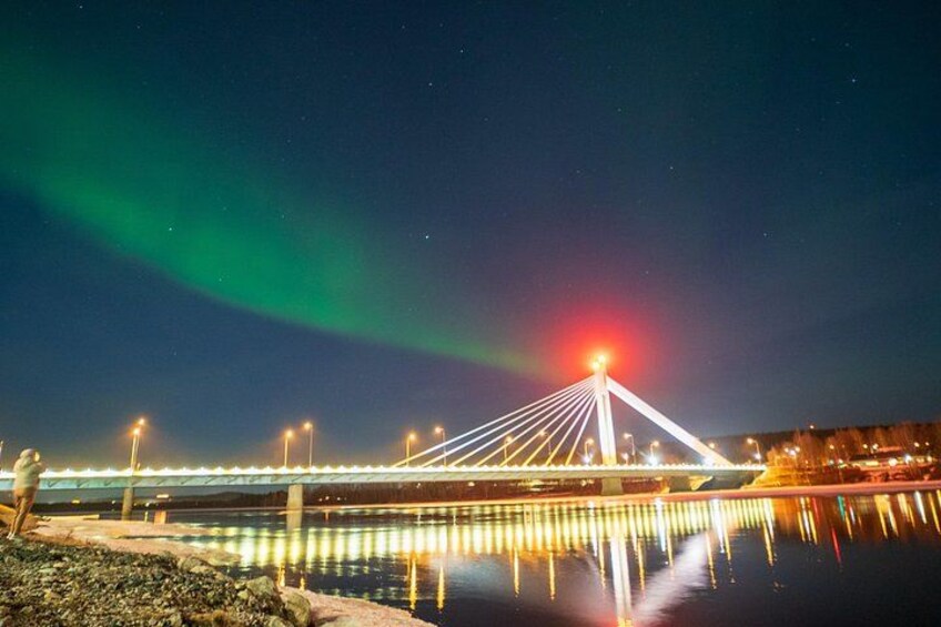 En bro med lys der spejler sig i vandet, et grønt nordlys og en by silhuet i baggrunden. 2