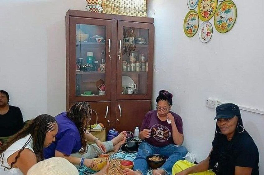 A group of people sitting on the floor in a room with a cabinet and decorative plates on the wall. 5