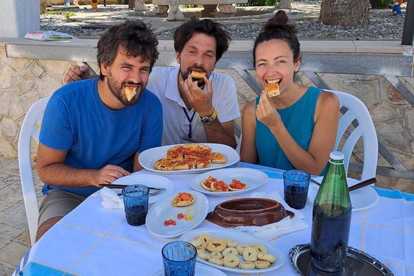 Trois personnes sont assises à une table en plein air, devant des assiettes remplies de nourriture, notamment des pizzas et des plats de pâtes, ainsi qu’une bouteille d’eau. 3