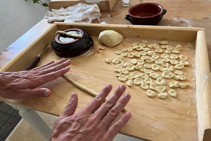 Des mains façonnent la pâte sur une surface en bois, avec un rouleau à pâtisserie et un bol d’eau à proximité. 1