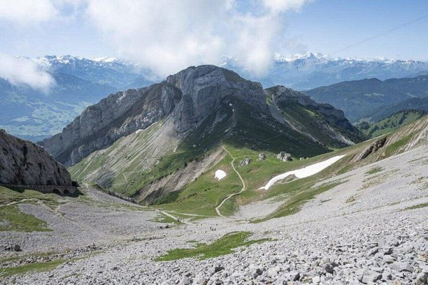 Paesaggio montuoso con terreno roccioso, un sentiero tortuoso e cime innevate in lontananza. 5