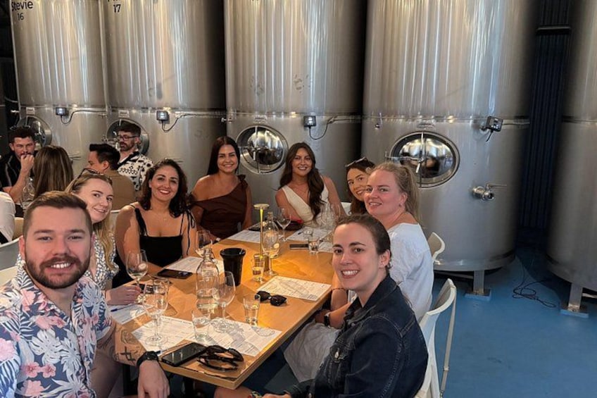 A group of people seated at a table in a winery with stainless steel tanks in the background. 1